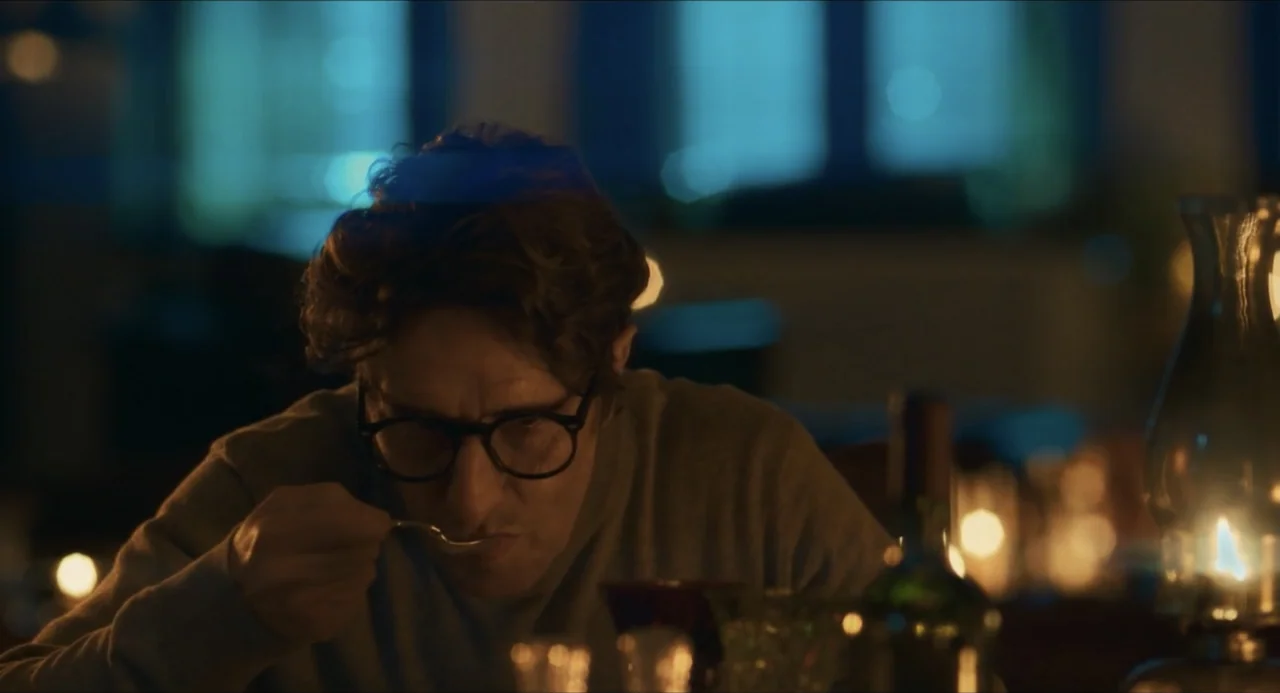 Echoes Of A Crime (2025) movie still: medium — A dimly lit room features a table set for dinner with candles and wine. A man wearing glasses sits at…
