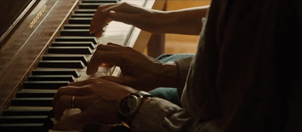 The Road (2009) movie still: closeup — A close view shows four hands playing a wood-grain Baldwin upright piano. Two individuals reach across the…