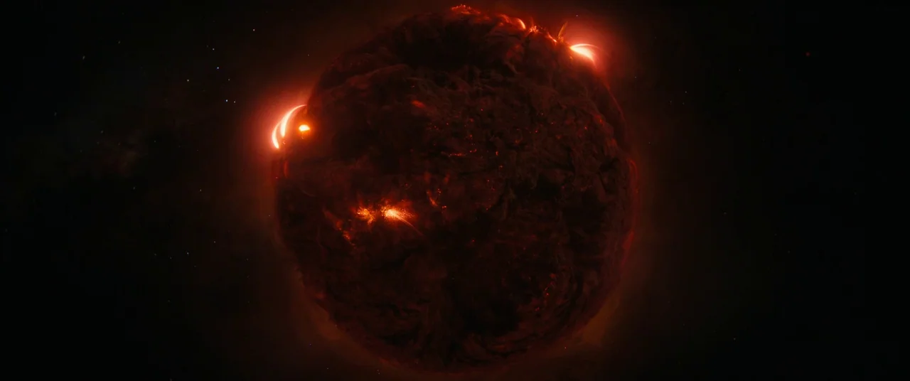 The Marvels (2023) movie still: establishing — A dark, textured planetoid drifts through the vacuum of deep space. Glowing red magma erupts from its…