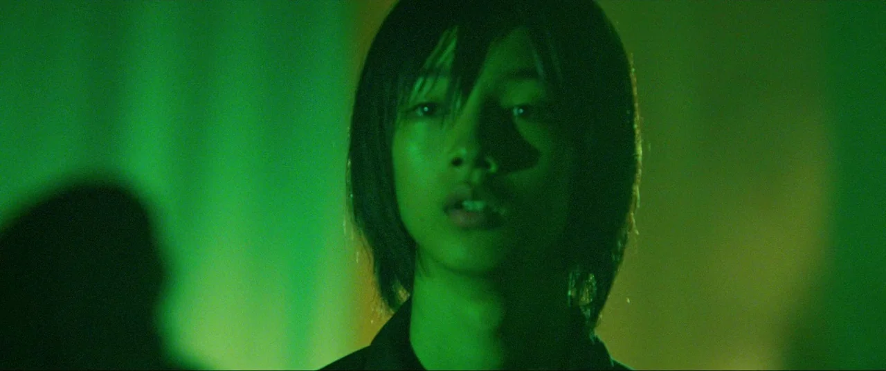 The World of Kanako (2014) movie still: closeup — A dark indoor environment is illuminated by an intense green light source. A young man with dark…