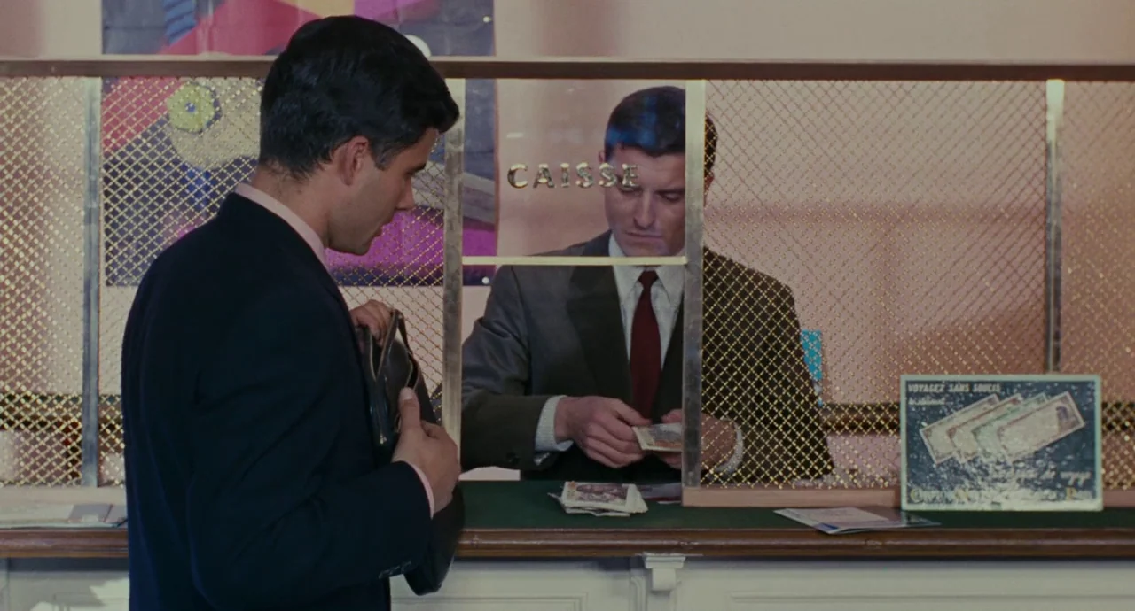 The Umbrellas Of Cherbourg (1964) movie still: medium — A man in a suit stands at a bank counter behind a brass wire mesh window. The bank teller…