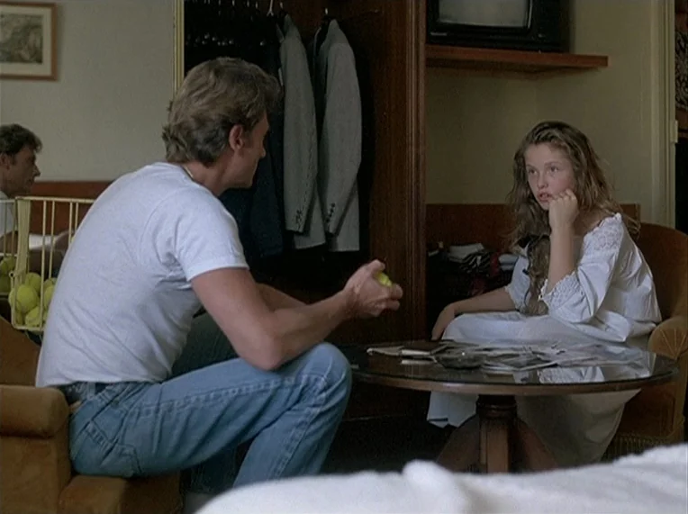 Detective (1985) movie still: medium — A hotel room with dark wood furniture and a glass topped coffee table. The man sits on a chair facing a girl…