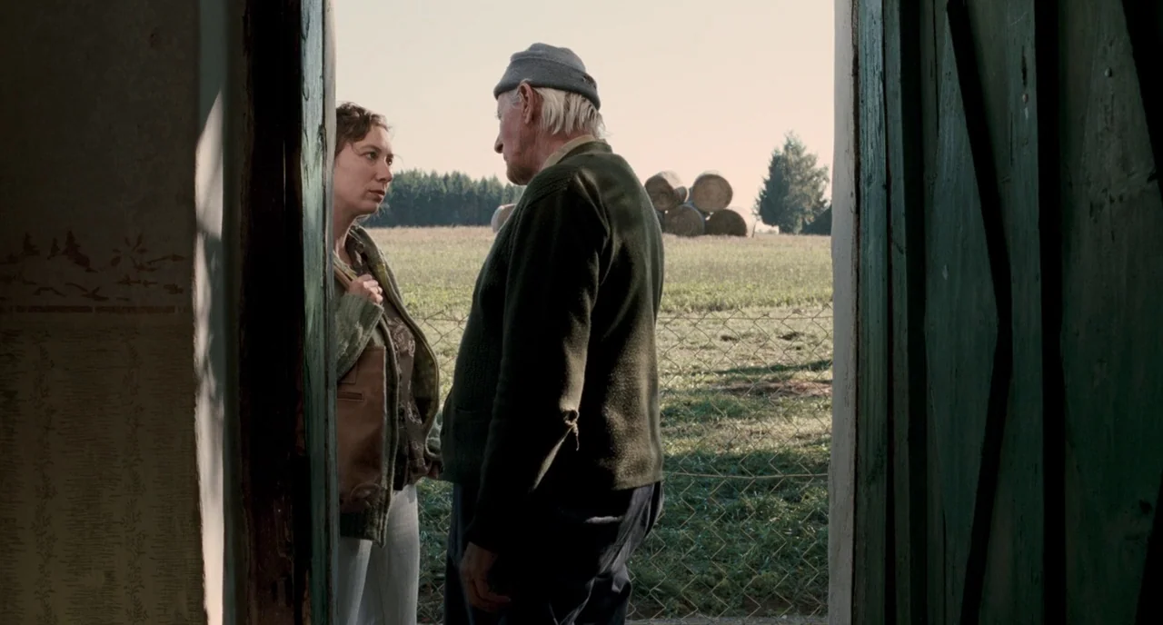 Revanche (2008) movie still: medium — A woman and an elderly man stand in the doorway of a rustic building. Beyond them is a rural landscape…