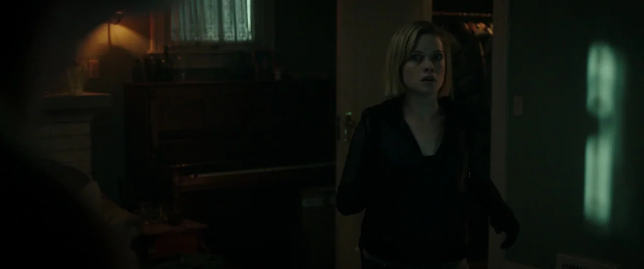 Don’t Breathe (2016) movie still: medium — A dimly lit room with muted teal walls and a dark upright piano against the wall. A woman in a black…