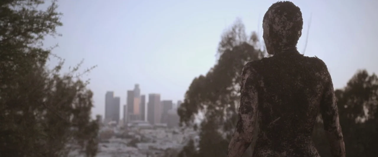 Starry Eyes (2014) movie still: medium — A figure covered in mud stands on a hillside overlooking a city skyline. The cityscape features multiple…