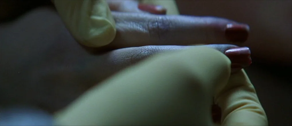 Insomnia (2002) movie still: closeup — A close view of a human hand being inspected by yellow latex gloves. The subject has dark red nail polish on…