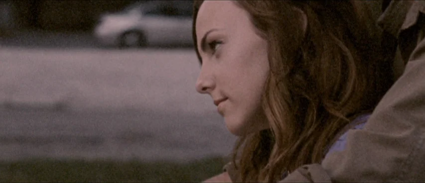 This Movie Is Broken (2010) movie still: closeup — An outdoor space at dusk with an asphalt surface visible in the background. A woman in a collared…