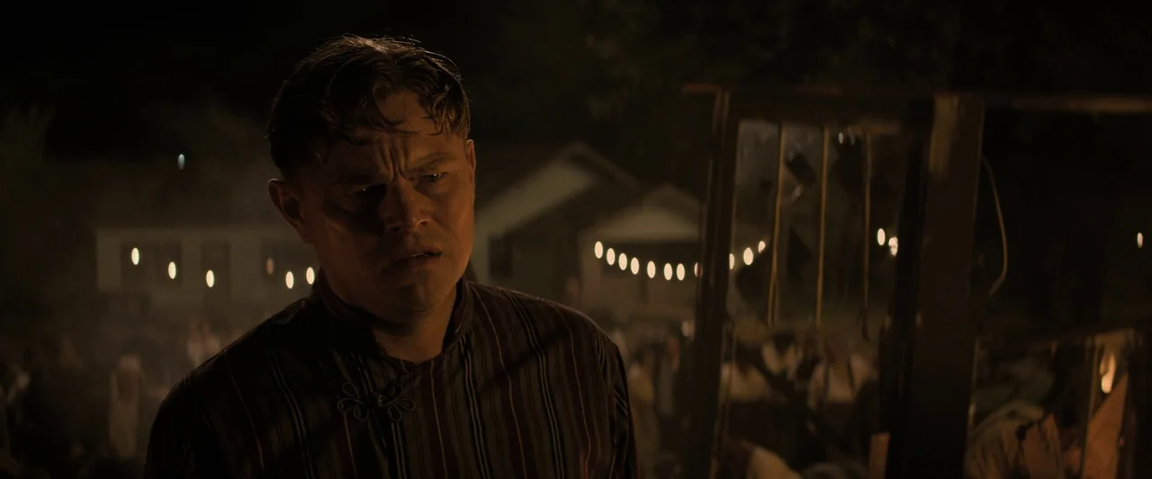 Killers of the Flower Moon (2023) movie still: medium — A night scene shows a man in a striped shirt standing at an outdoor event. Blurred string…