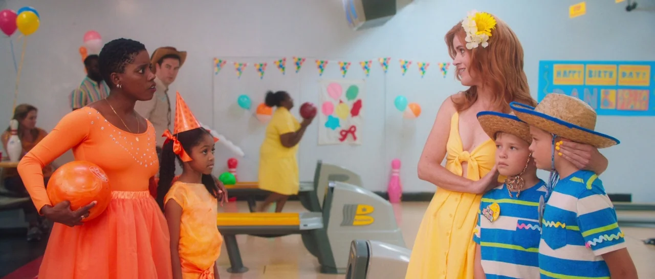 Greener Grass (2019) movie still: wide — A brightly lit, decorated venue features festive balloons and children's party banners. Two mothers stand…