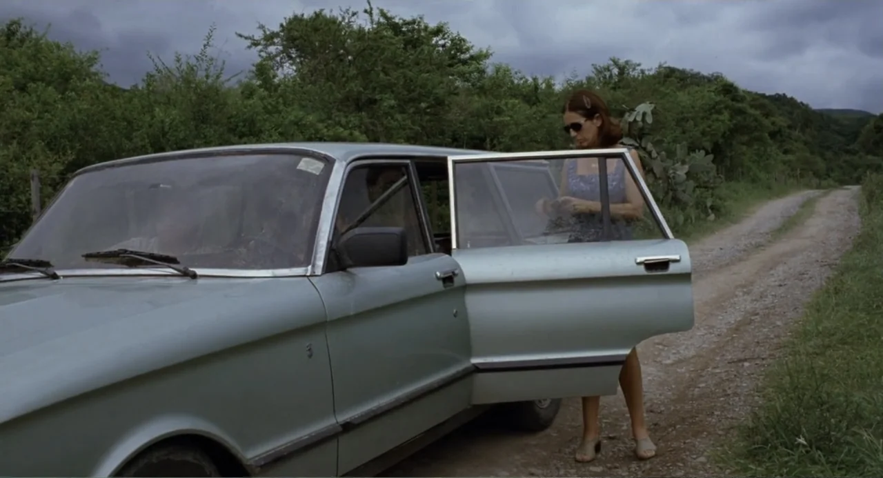 La Ciénaga (2001) movie still: fullbody — A silver sedan sits parked on a narrow dirt path bordered by dense green vegetation. A woman stands beside…