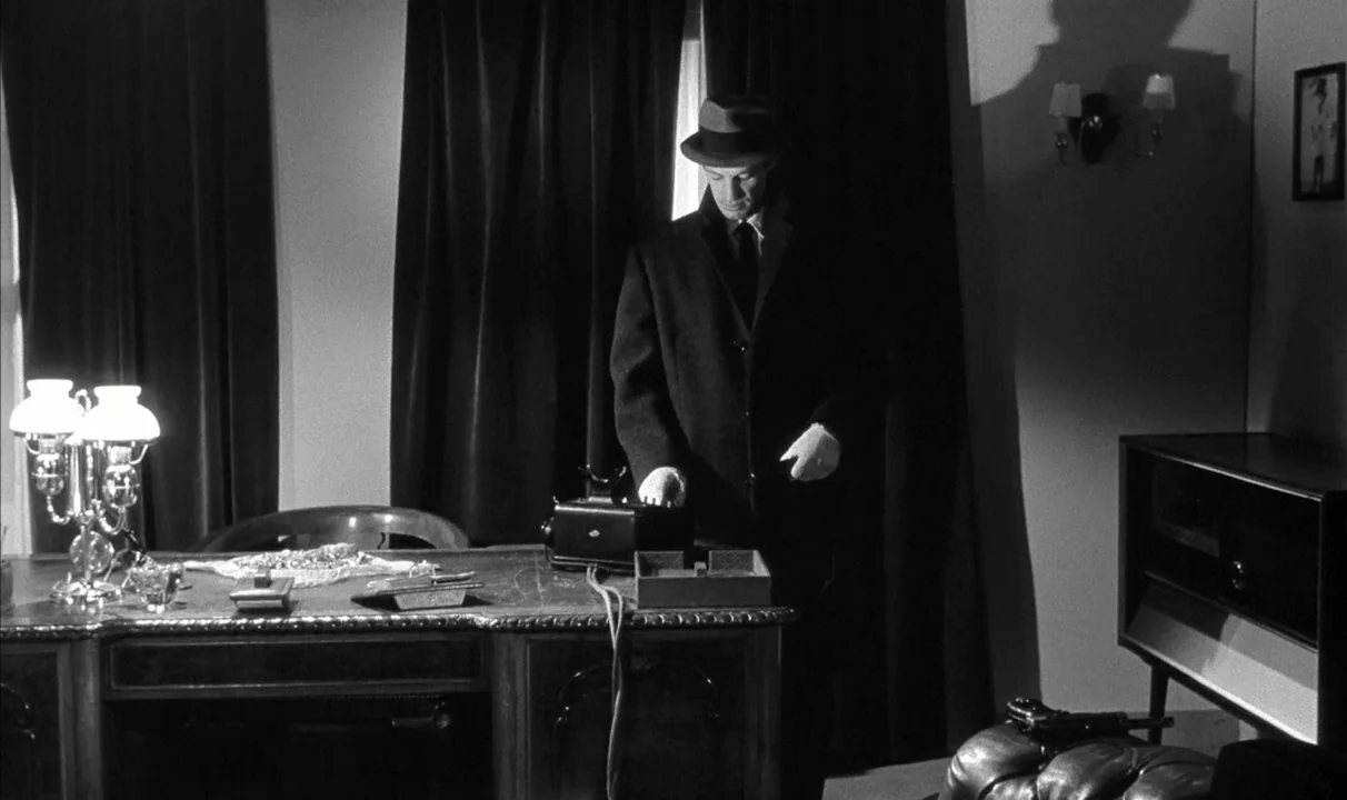 Le Doulos (1962) movie still: medium — A dimly lit 1950s study features heavy dark curtains and a large antique desk. A man in a hat stands over the…