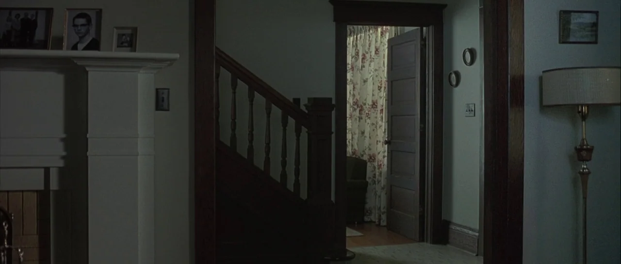 Capote (2005) movie still: wide — A dark, dimly lit domestic space features a staircase and a doorway leading inward. Floral curtains hang in the…