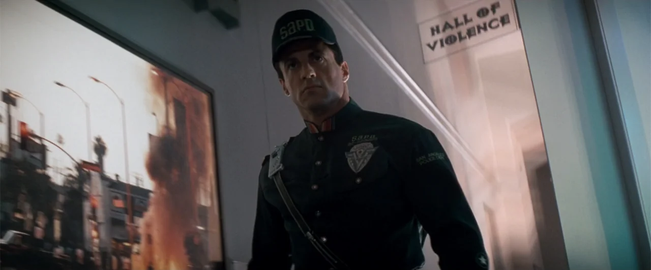 Demolition Man (1993) movie still: medium — An interior hallway features a wall-mounted digital display showing an urban fire scene. A man in a dark…