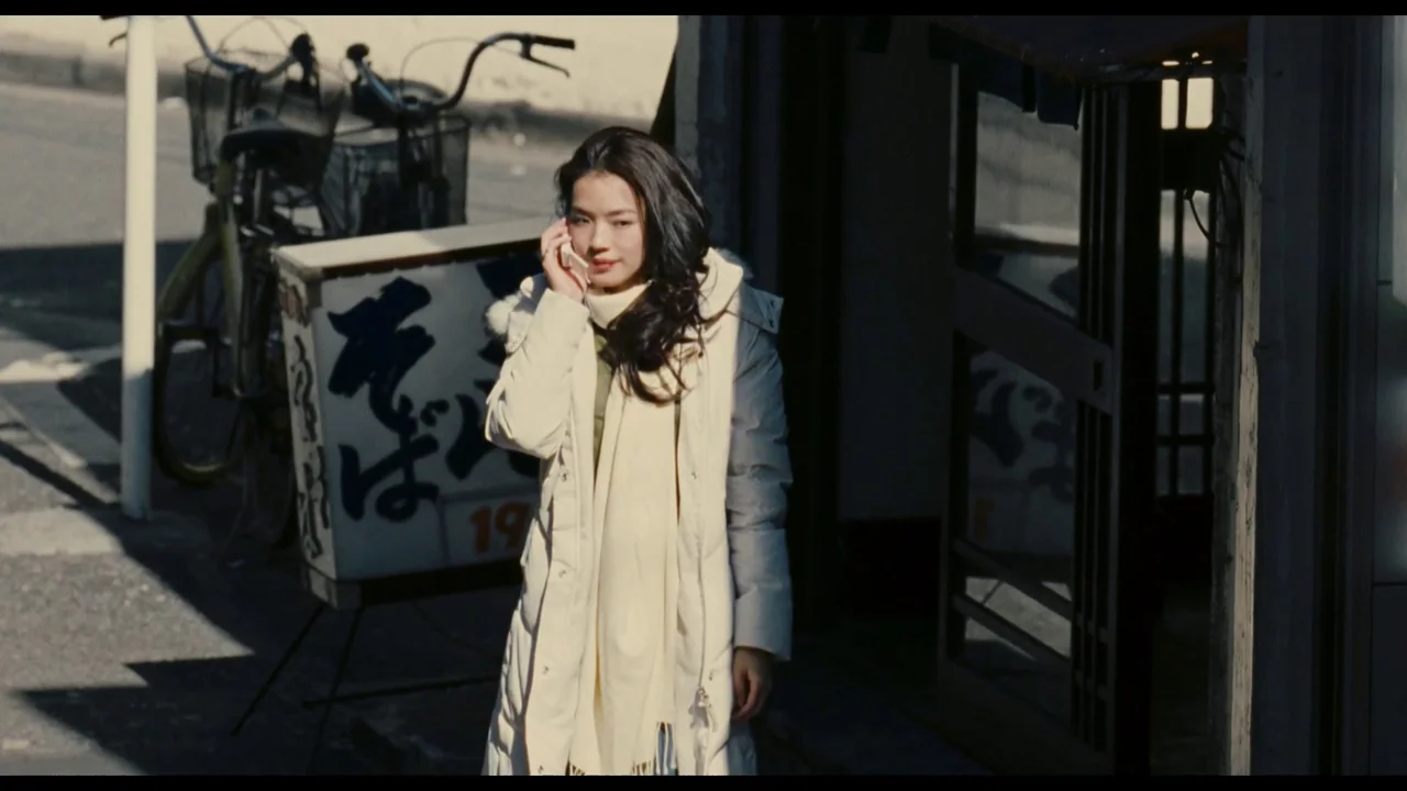 Millennium Mambo (2001) movie still: medium — An outdoor urban sidewalk beside a wooden storefront with a nearby parked bicycle. A young woman in a…