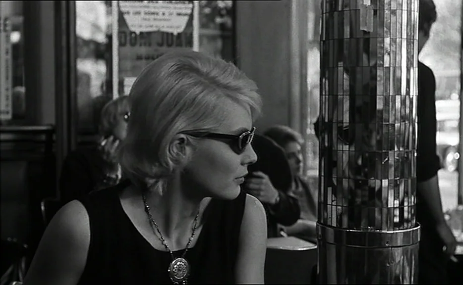 Cléo from 5 to 7 movie still: medium — A cafe interior with a reflective mirrored pillar in the foreground. A blonde woman in sunglasses sits at a…