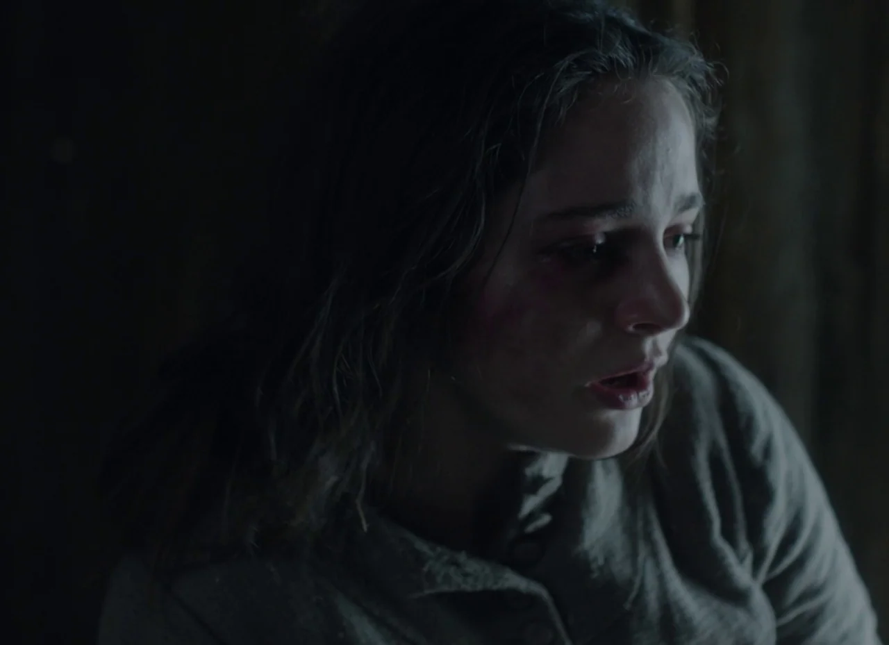 The Nightingale (2018) movie still: closeup — A dimly lit, dark space surrounds a woman in a textured top. The woman has disheveled hair and appears…