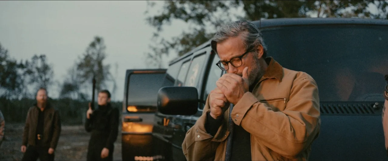 Ambush (2020) movie still: medium — A man stands in a dirt field near a parked dark van. He focuses on lighting a cigarette while two armed figures…