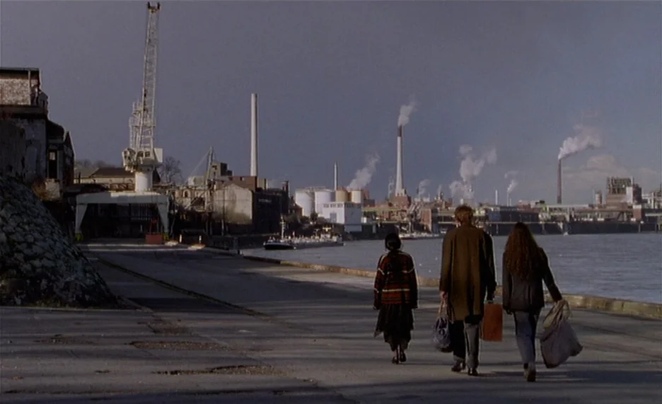 Pola X (1999) movie still: wide — An industrial waterfront area features a concrete quay bordered by a harbor and factory buildings. Three people…