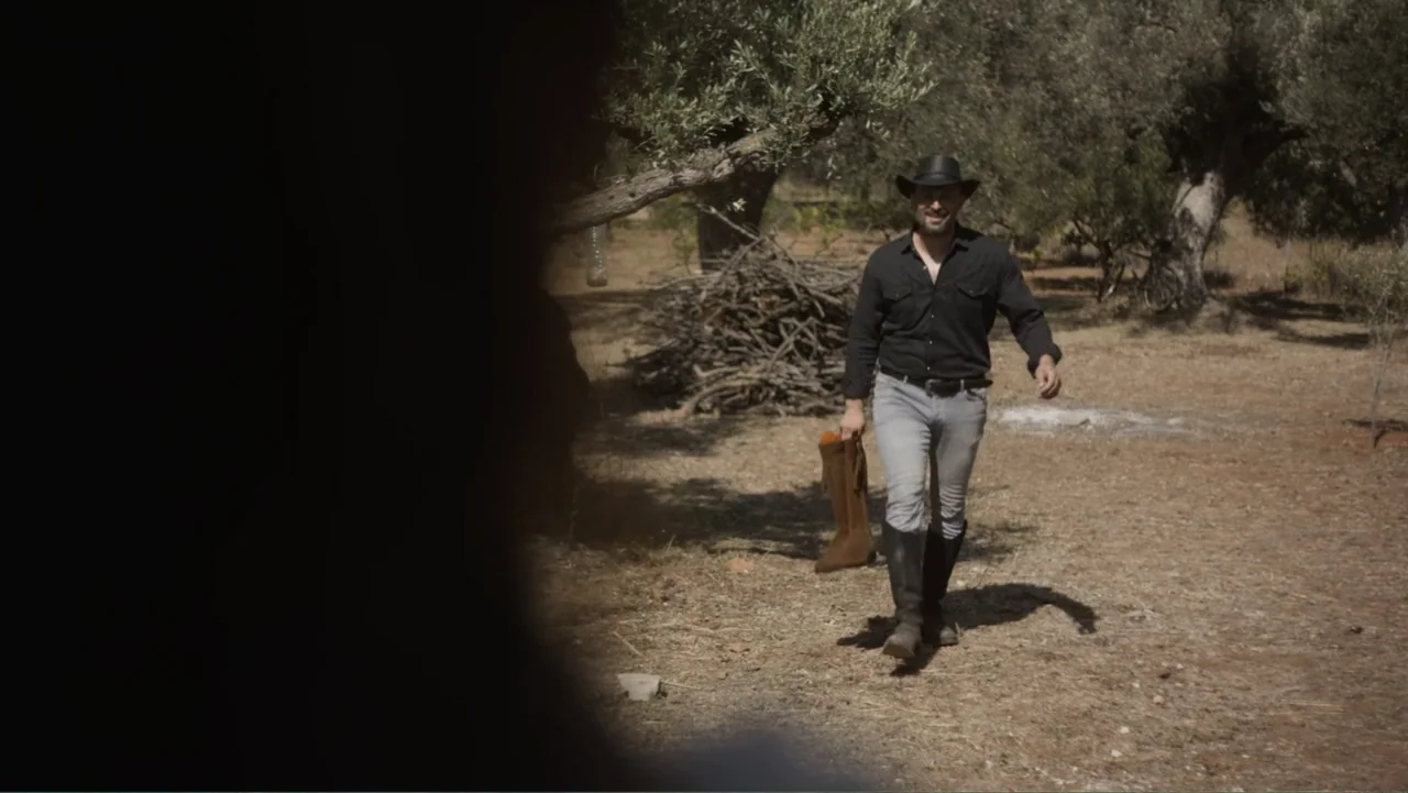 Hot Milk (2025) movie still: fullbody — A dry field with sparse olive trees and piles of gathered wooden branches. A man in a black shirt and hat…