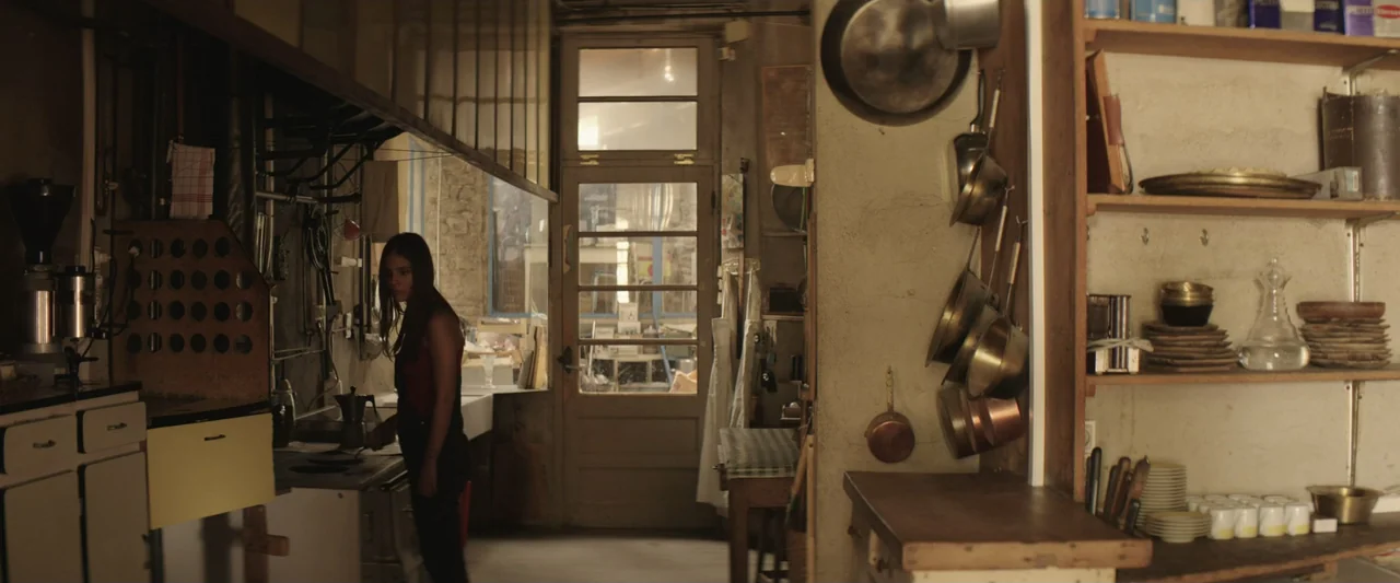 The Only One (2021) movie still: medium — A cluttered kitchen features copper pots hanging on a wall and open shelving units. A young woman in dark…