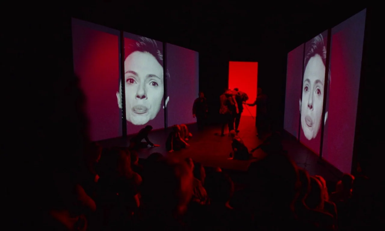 Marriage Story movie still: wide — A dark theater stage features large vertical screens projecting a woman's close-up face. Several figures move…