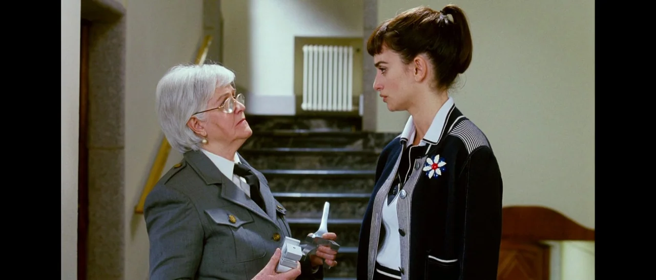Broken Embraces (2009) movie still: medium — A woman in a grey uniform stands in a residential hallway near stairs. She faces a younger woman with…