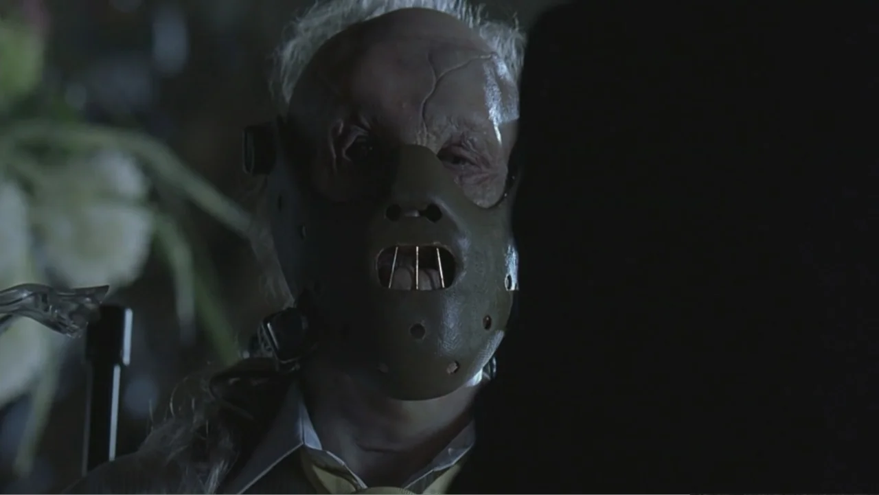 Hannibal (2001) movie still: closeup — An interior space dimly lit, focusing on a man wearing a brown leather muzzle mask. The man faces forward,…