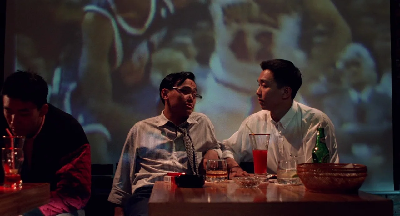 A Confucian Confusion (1994) movie still: medium — A dimly lit bar with a wooden table cluttered with drinks and ashtrays. Two men sit in…