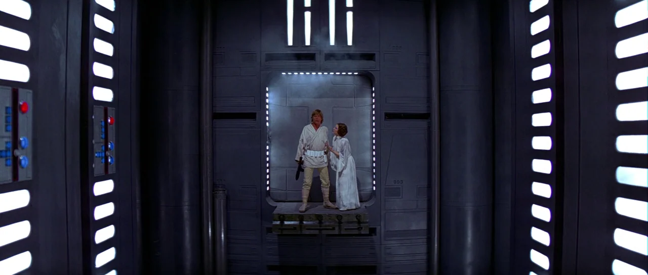 Star Wars (1977) movie still: fullbody — A futuristic corridor featuring grey, paneled walls and vertical strips of recessed lighting. A man and a…