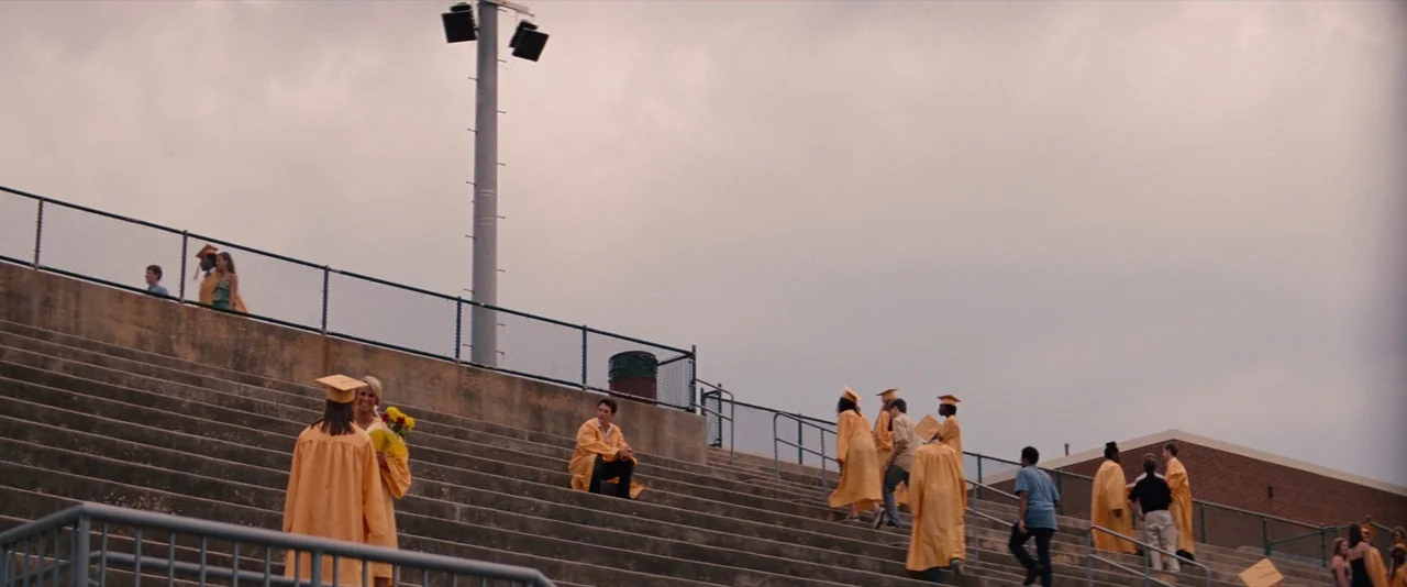 The Spectacular Now (2013) movie still: wide — Concrete stadium bleachers feature a group of graduates wearing yellow robes under cloudy skies. A…