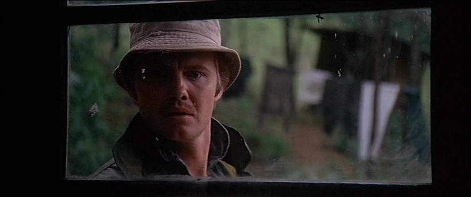 Deliverance (1972) movie still: closeup — A man looks through a window from a wooded exterior toward an indoor space. Laundry hangs on a clothesline…