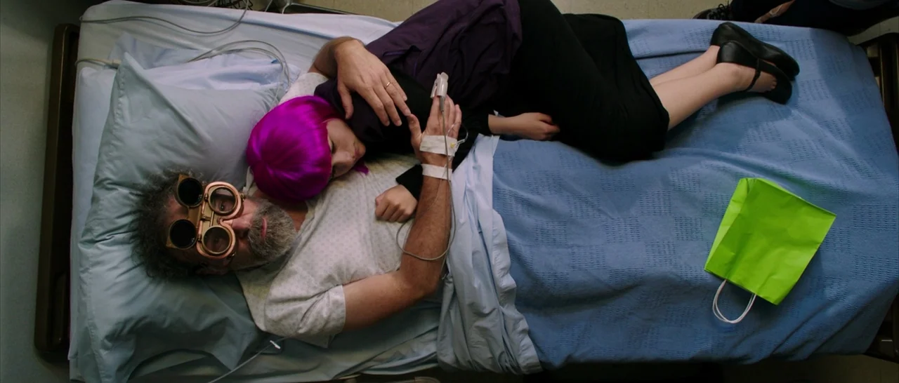 Wish I Was Here (2014) movie still: wide — A top-down view of a hospital bed with blue sheets and white pillows. A woman with bright pink hair rests…