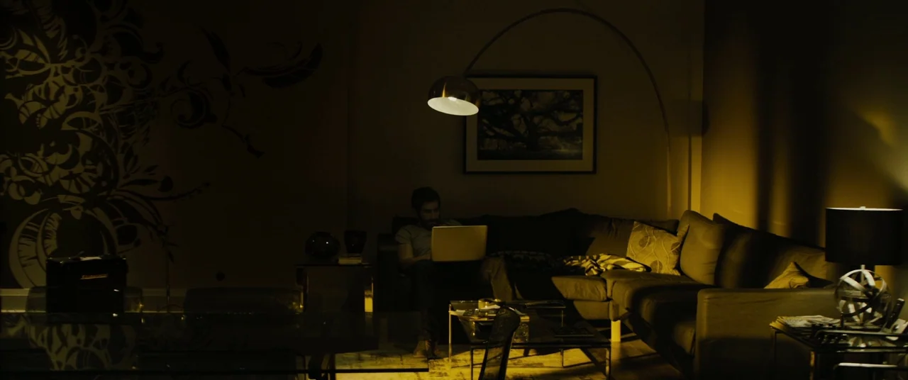 Enemy (2013) movie still: wide — A dark, dimly lit space features a man sitting on a large sectional couch. He operates a laptop while positioned…