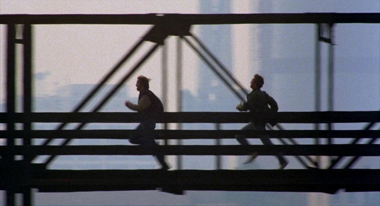 To Live and Die in L.A. (1985) movie still: wide — A metal truss bridge spans the frame against a pale, bright overcast sky. Two men in dark…