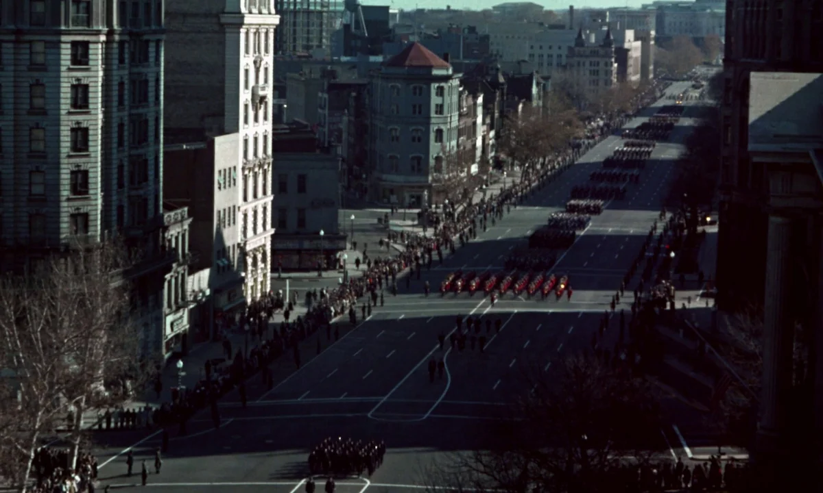 Jackie (2016) movie still: establishing — An aerial perspective shows a wide urban boulevard lined with tall, classic city buildings. Rows of…