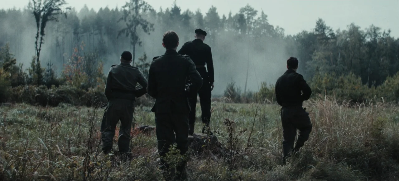 The Tank (2025) movie still: fullbody — A group of men stand in a misty, overgrown field bordering a dense forest. They watch a patch of smoke…