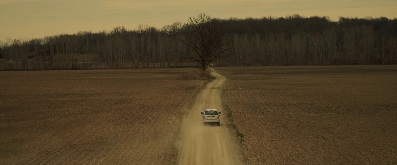You’re Next (2013) movie still: wide — An empty dirt road cuts through vast, flat, fallow fields toward a tree line. A white SUV travels down the…