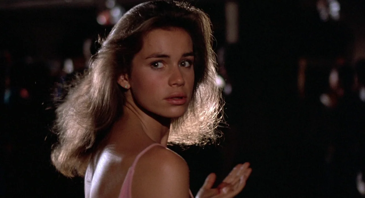 Breathless (1983) movie still: closeup — A dark, out-of-focus background suggests a busy interior space like a night club. A young woman with long…