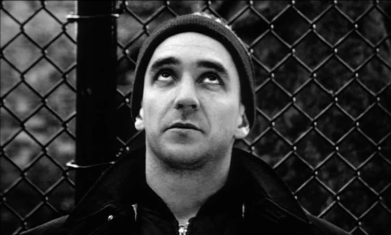 Pi (1998) movie still: closeup — A metal chain link fence serves as a backdrop for the close-up shot. A man in a dark knit cap and jacket looks up…