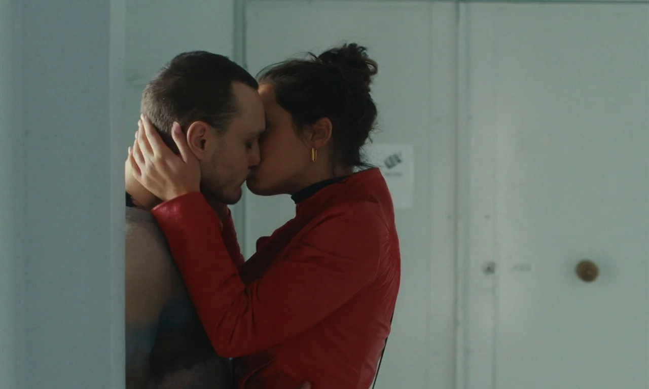 Passages (2023) movie still: medium — A dimly lit residential hallway with white doors and a simple door knob. A young man and a woman are kissing…