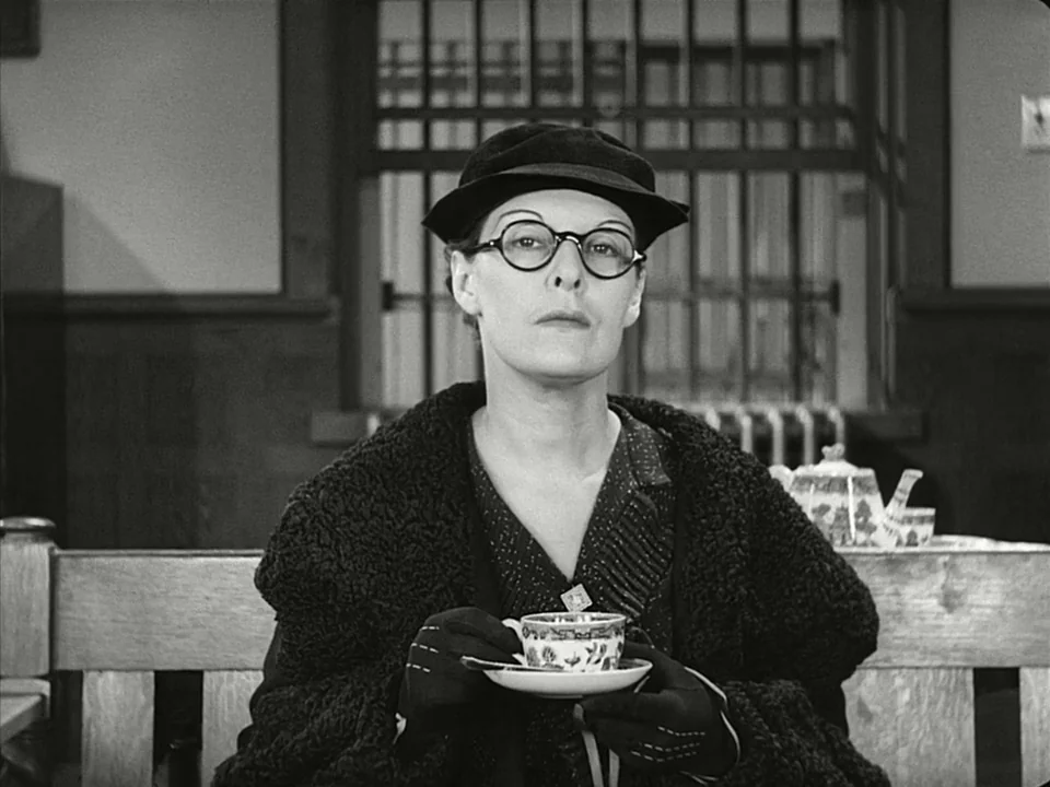 Modern Times (1936) movie still: medium — A woman sits at a table in a dimly lit establishment with metal window bars. She holds a small teacup and…