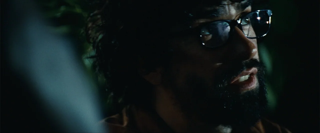 Battleship (2012) movie still: closeup — A man sits in a dimly lit, shadowed space with dark foliage blurred in the background. The man with dark…