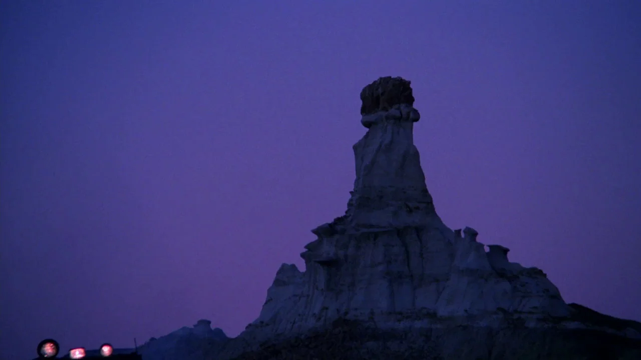 Sorcerer (1977) movie still: wide — A tall, eroded sandstone hoodoo stands against a deep violet twilight sky. The rock formation occupies the…
