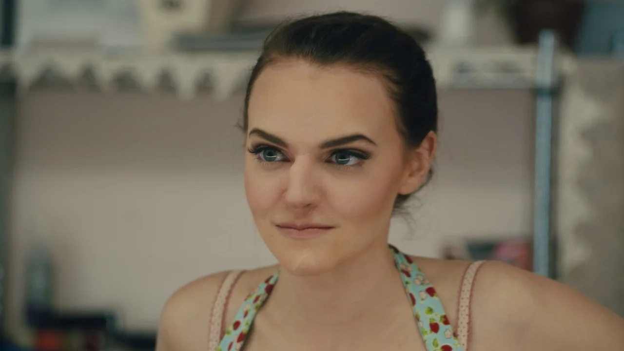 Cam (2018) movie still: closeup — A workspace with a wire rack shelf is visible behind the subject. A woman looks off-camera while wearing a…