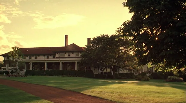 The Virgin Suicides (1999) movie still: establishing — A sprawling historic mansion with a red roof sits behind a manicured green lawn. A dirt path…