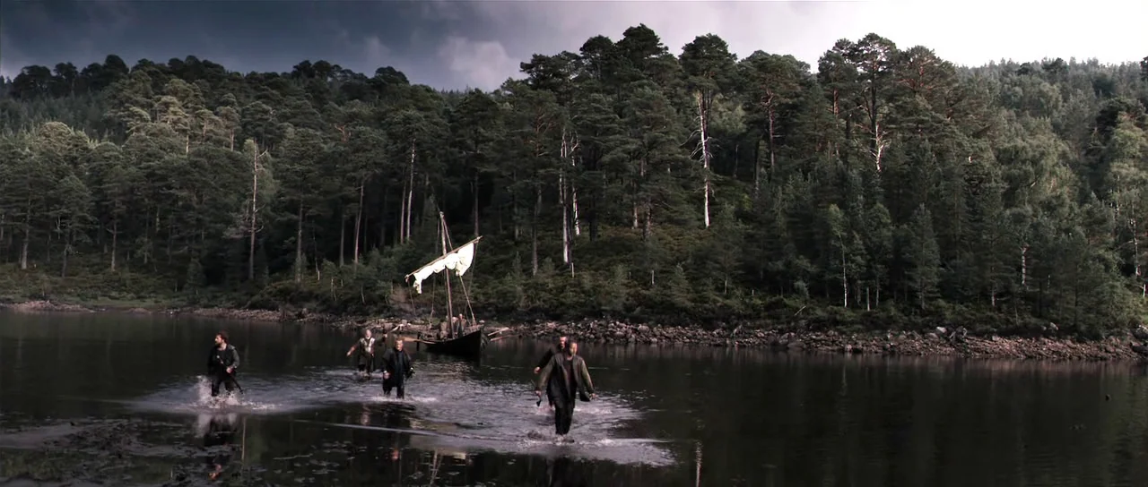 Valhalla Rising (2009) movie still: wide — A group of men wade through the shallow water of a dark lake. A small wooden boat with a simple sail sits…