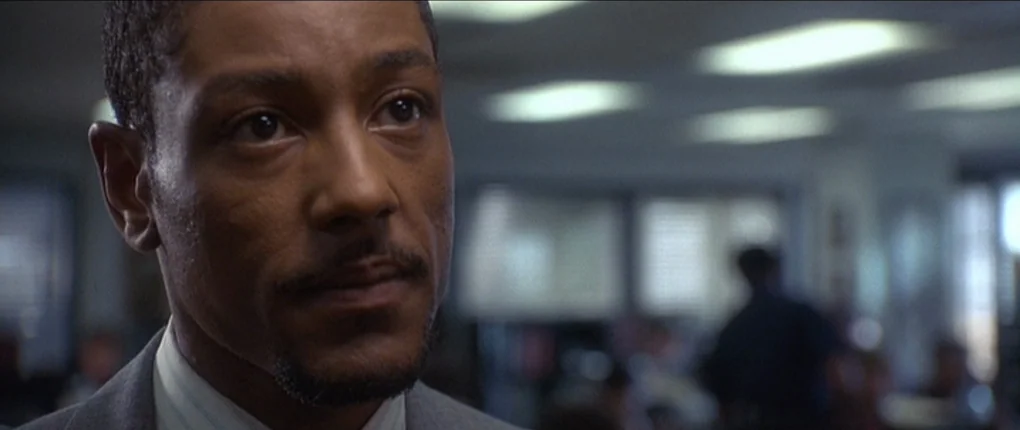 The Usual Suspects movie still: closeup — A man stands in a dimly lit, blurred professional environment with fluorescent ceiling lights. The subject…