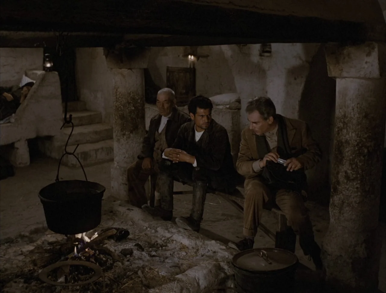 Christ Stopped at Eboli (1979) movie still: medium — A subterranean room with rough stone pillars and a dirt floor dimly lit by fire. Three men sit…