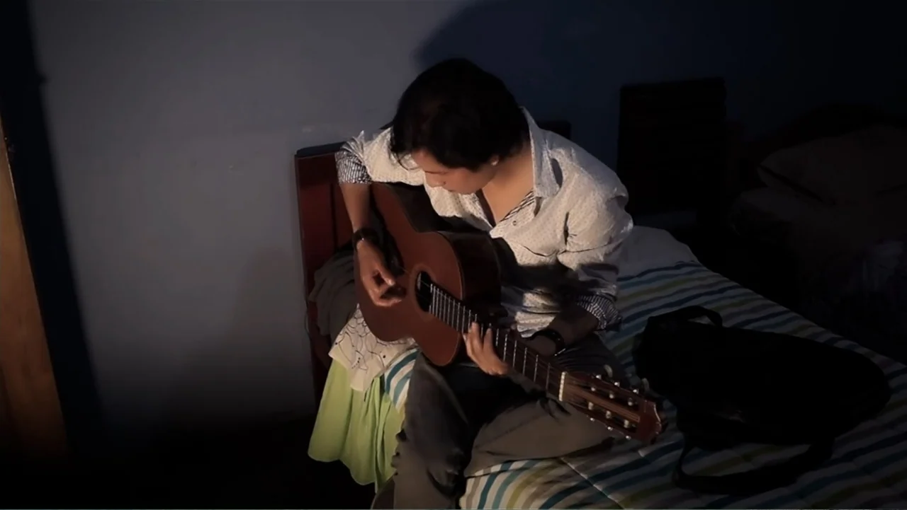 El Duende Sátiro (2016) movie still: medium — A dim room contains a wooden headboard and a striped blanket on a bed. A young man sits on the bed…