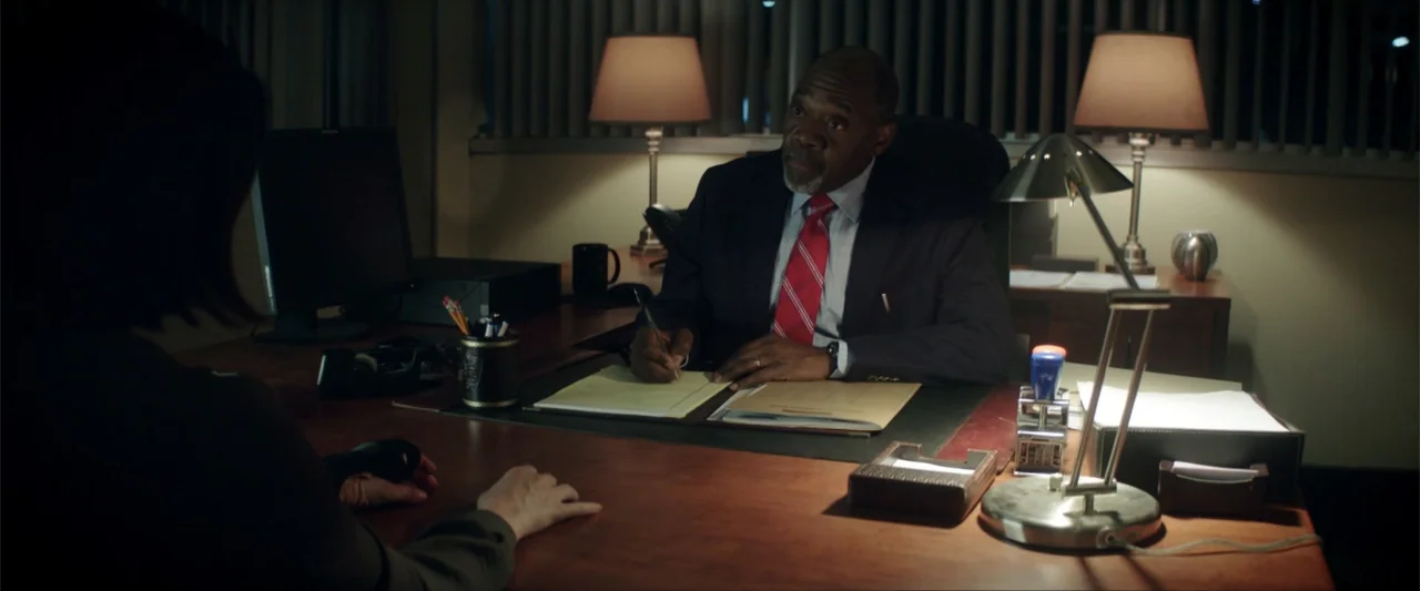 Truth or Dare (2018) movie still: medium — A dimly lit office features a large wooden desk with lamps and paperwork. An older man in a suit sits…